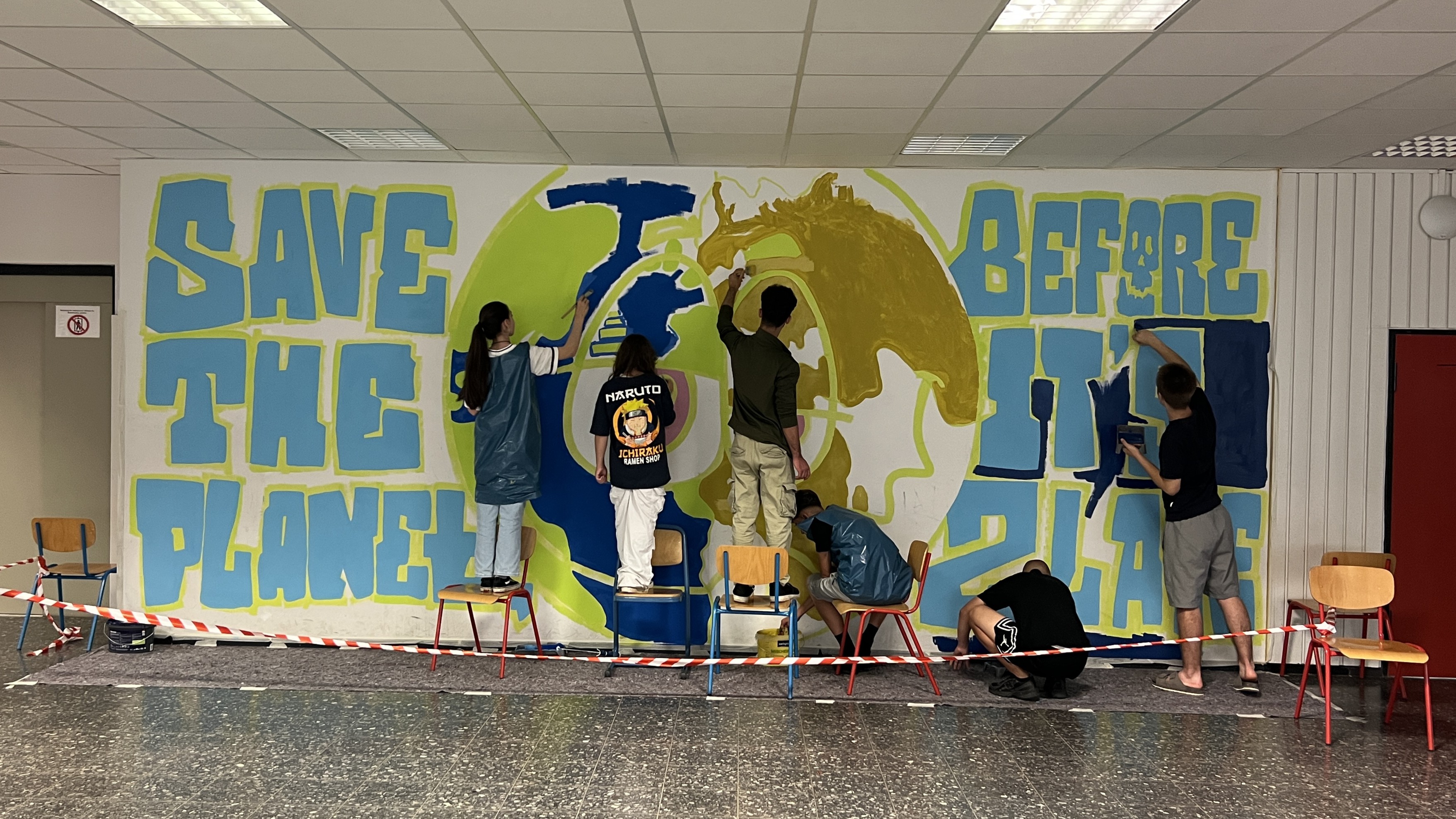 Mehrere Menschen malen eine Wand in einem Gebäude an. Auf der Wand ist eine Erdkugel und der Schriftzug 